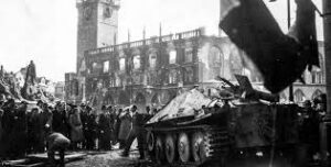 Black-and-white photo of a damaged tank in a city square with a burned building and crowd of civilians nearby.